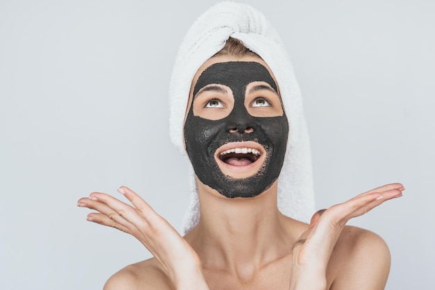 Studio shot of happy young woman with facial cosmetic black organic clay mask on her face looks up to advertising text wears white towel on hair isolated over white wall Healthy beauty concept