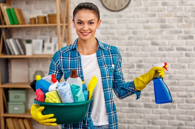 Студийный снимок домработницы. Комната уборки красивой женщины. Женщина в перчатках, улыбается и держит миску, полную бутылок с дезинфицирующим средством