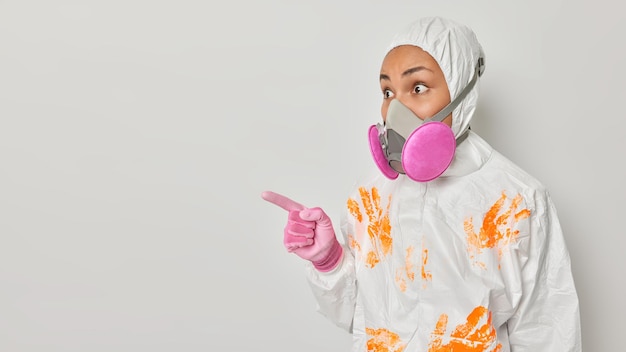 Studio shot of impressed female ecological activist tries to prevent exhausting toxic gases into air points index finger away on copy space wears protective suit and gas mask isolated over white wall