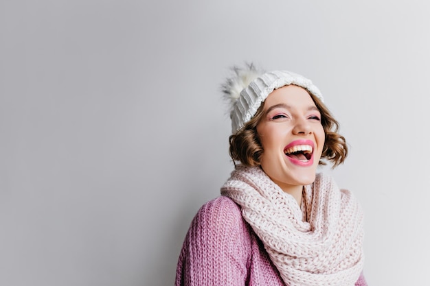 Studio shot of laughing winsome female model in trendy knitted hat. Romantic girl in woolen scarf smiling on light background.