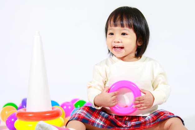 Студийный снимок маленькой милой азиатской модели дочери девочки в повседневной одежде, сидящей на полу и играющей с красочными пластиковыми кольцами, строительной пирамидой, игрушкой и маленькими шариками на белом фоне.