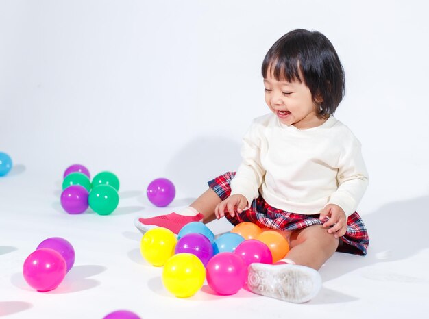 Studio shot of little cute short black hair Asian baby girl daughter model in casual plaid skirt sitting on floor smiling laughing playing with colorful round balls toy alone on white background.