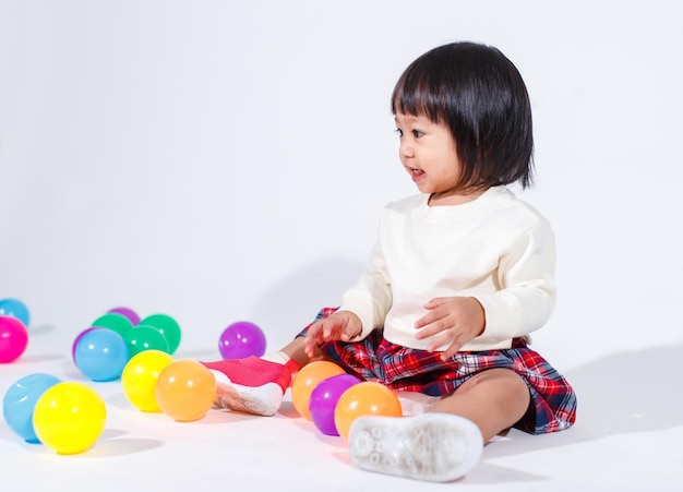 Studio shot of little cute short black hair Asian baby girl daughter model in casual plaid skirt sitting on floor smiling laughing playing with colorful round balls toy alone on white background.