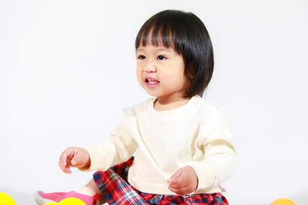 Studio shot of little cute short black hair Asian baby girl daughter model in casual plaid skirt sitting on floor smiling laughing playing with colorful round balls toy alone on white background.