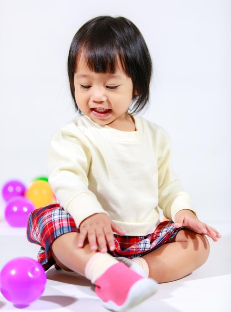 Studio shot of little cute short black hair Asian baby girl daughter model in casual plaid skirt sitting on floor smiling laughing playing with colorful round balls toy alone on white background.