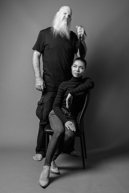 Studio shot of mature bearded bald man and young beautiful asian woman together against gray wall in black and white