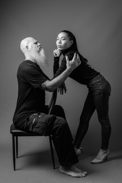 Studio shot of mature bearded bald man and young beautiful Asian woman together against gray wall in black and white