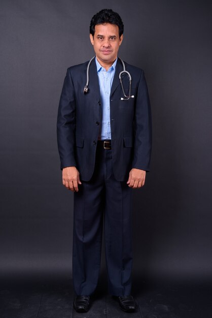 Studio shot of mature handsome Indian businessman in suit as doctor with stethoscope against black background