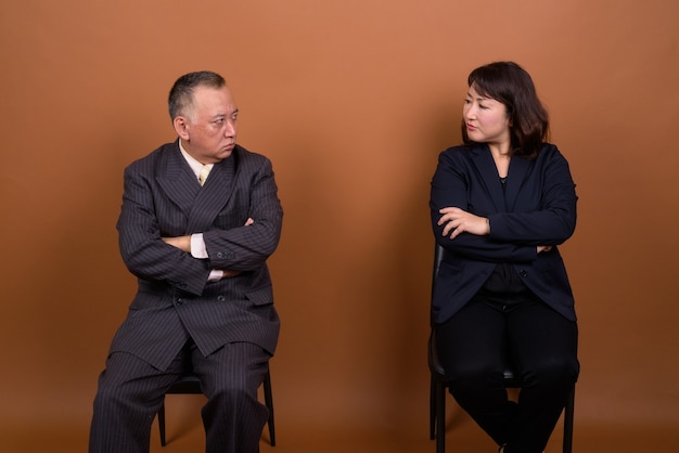 Studio shot of mature Japanese businessman and mature Japanese businesswoman together against brown background