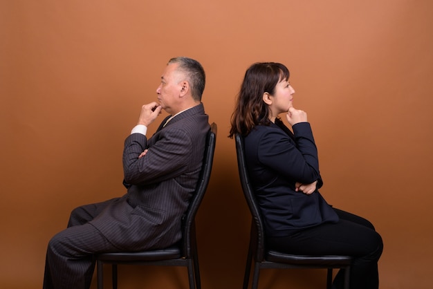 Studio shot of mature Japanese businessman and mature Japanese businesswoman together against brown background