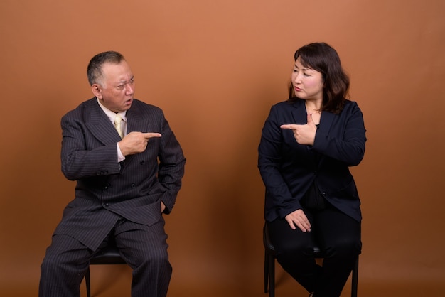 Studio shot of mature Japanese businessman and mature Japanese businesswoman together against brown background