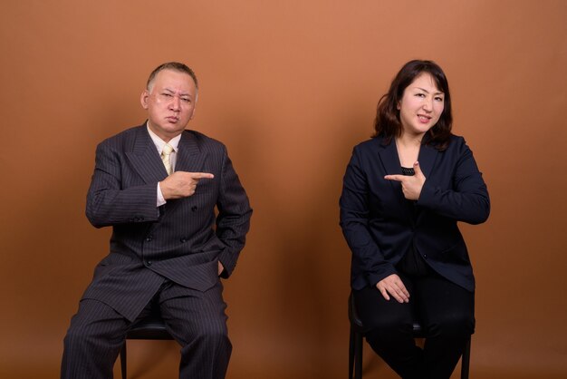 Studio shot of mature Japanese businessman and mature Japanese businesswoman together against brown background