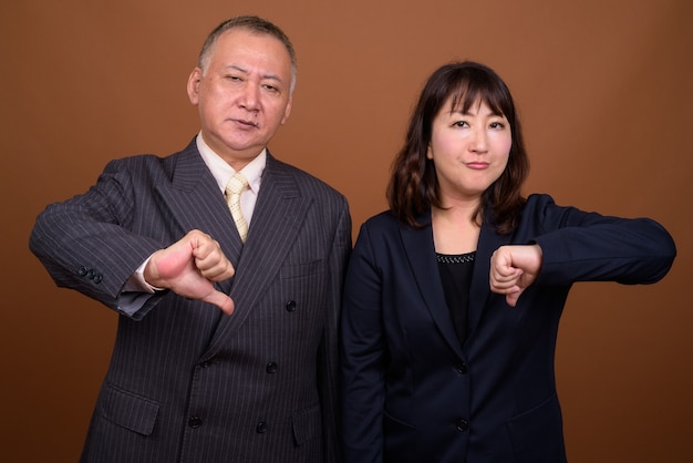 Studio shot of mature Japanese businessman and mature Japanese businesswoman together against brown background