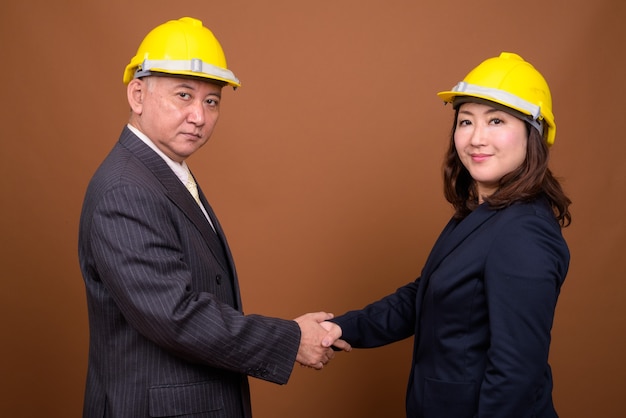 Studio shot of mature Japanese businessman and mature Japanese businesswoman wearing hardhat together against brown background