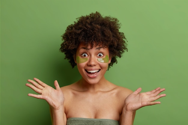 写真 陽気な女性のスタジオショットは手のひらを広げ、何かに喜んで反応します目の下にコラーゲンパッチを適用し、緑のスタジオの壁に孤立した腫れやしわを減らします