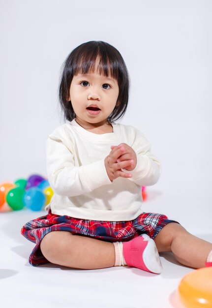 写真 白い背景の上のカラフルな丸いボールのおもちゃだけで遊んで笑って笑って笑って床に座っているカジュアルな格子縞のスカートで小さなかわいい短い黒髪アジアの女の赤ちゃん娘モデルのスタジオショット。