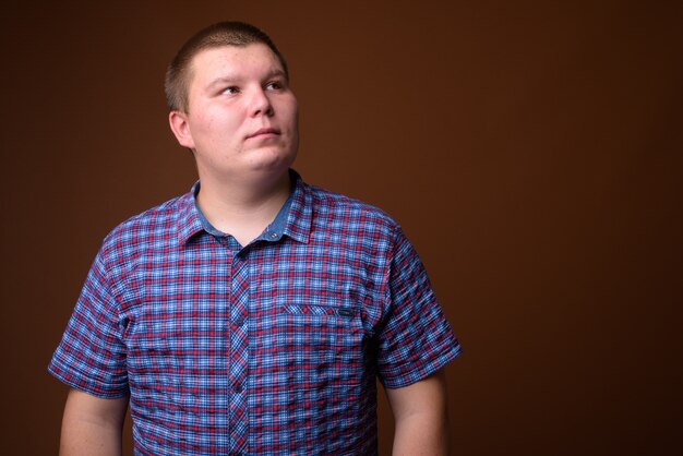 Photo studio shot of overweight young man wearing purple checkered shirt against brown background