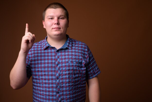 Studio shot of overweight young man wearing purple checkered shirt against brown background