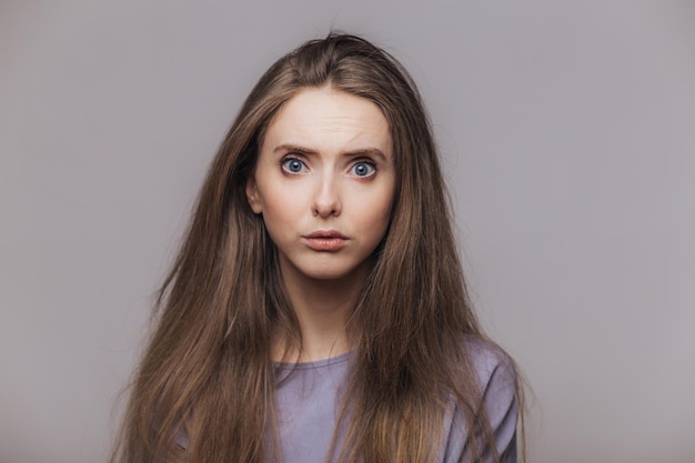 Studio shot of pleasant looking blue eyed female model with dark long hair has puzzled expression as hears shocking news from interlocutor isolated over grey background Facial expressions concept