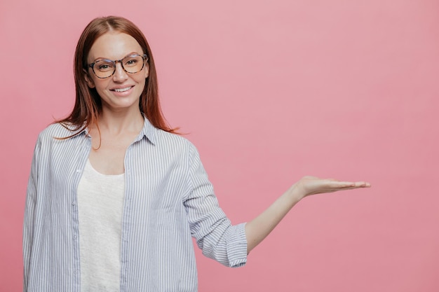 Studio shot of pleased Caucasian woman keeps palm raised wears optical glasses demonstrates something over copy space holds blank space has pleasant smile on face poses indoor over pink wall