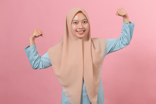 Photo studio shot of positive young asian muslim woman raises arms shows muscles pretends to be very strong and powerful smiles gently wears hijab isolated over pink background.