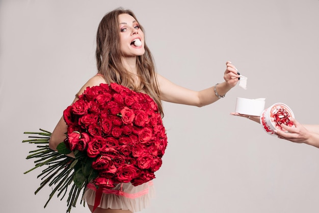 Studio shot of pretty brunette woman holding bouquet of gorgeous red roses She is eating sweet and taking one more candy from the box in anonymous female hands Isolate on white