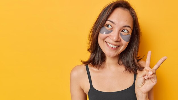 Studio shot of pretty dreamy woman with dark hair makes peace\
sign keeps fingers raised has satisfied expression dressed in black\
t shirt isolated over yellow background copy space for your\
advert