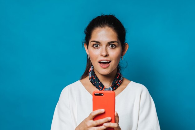 Studio shot of pretty woman stares at mobile phone, feels surprised, opens mouth, dressed in white blouse and neckerchief, isolated over blue background with blank space for your promotion