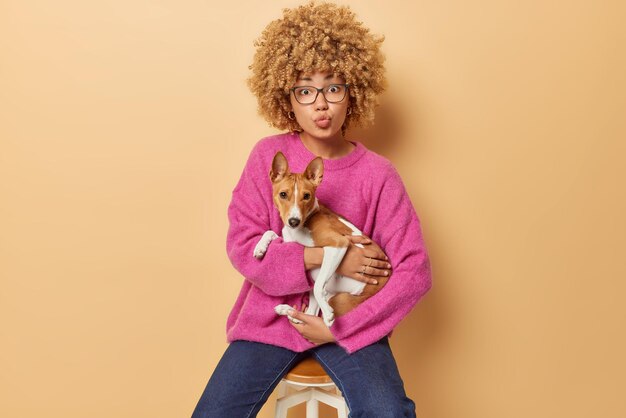 Studio shot of pretty young woman has curly hair keeps lips\
folded carries basenji dog dressed in pink jumper jeans poses on\
chair isolated over beige background going to have morning walk\
with pet