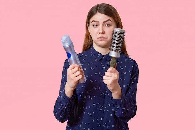 Studio shot of puzzled dark haired young woman holds hairspray and comb