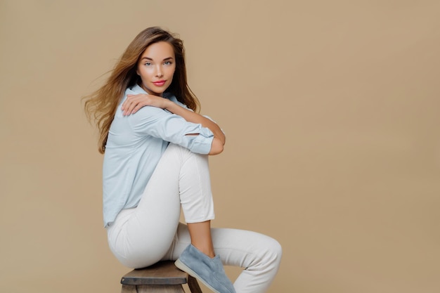 Studio shot of restful beautiful Caucasian woman sits on chair wears shirt white trousers and shoes keeps hands crossed over body has confident look at camera isolated over brown background