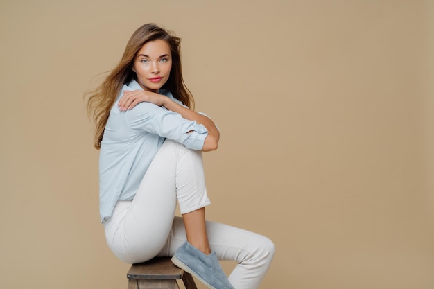 Studio shot of restful beautiful Caucasian woman sits on chair wears shirt white trousers and shoes keeps hands crossed over body has confident look at camera isolated over brown background