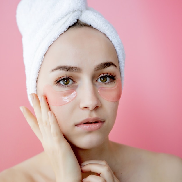 Studio shot of satisfied caucasian freckled woman wearing white towel on head, with collagen patches under eyes, standing naked against pink background. Skin care, cosmetic product concept