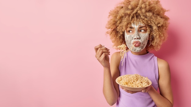 Studio shot of serious woman looks attentively on left side\
eats spaghetti applies beauty facial mask for skin moisturising\
dressed in casual t shirt poses against rosy background mock up\
space