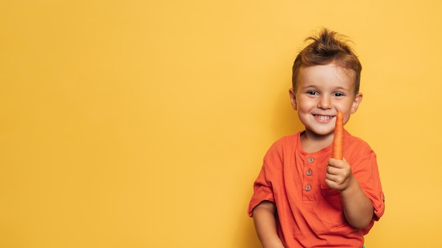 Studio shot of a smiling boy holding a fresh carrot on a yellow background The concept of healthy baby food carotene A place for your text