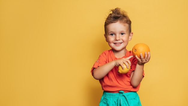 黄色の背景に新鮮なレモンとオレンジを保持している笑顔の少年のスタジオショット。健康的な離乳食、ビタミンCの概念。あなたのテキストのための場所。