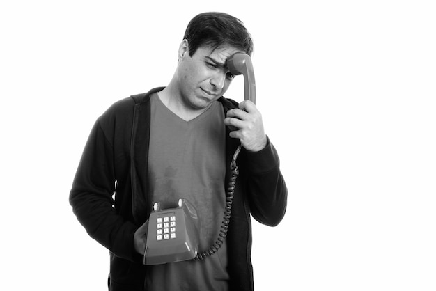 Studio shot of stressed young Persian man looking tired while holding old telephone on head isolated