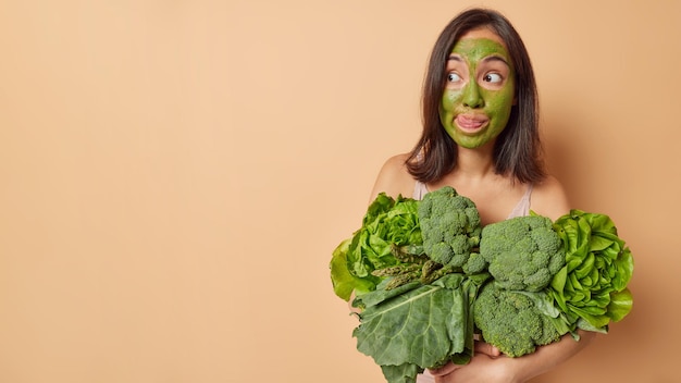 Studio shot of surprised dark haired young woman licks lips\
with tongue focused away applies green facial mask to remove fine\
lines and having smooth skin carries vegetales keeps to healthy\
diet