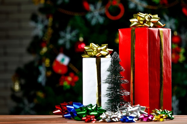 Photo studio shot of tall gift boxes with shiny golden ribbon placed on wooden table with colorful glossy bow tie in front decorative beautiful christmas eve pine tree in blurred background.