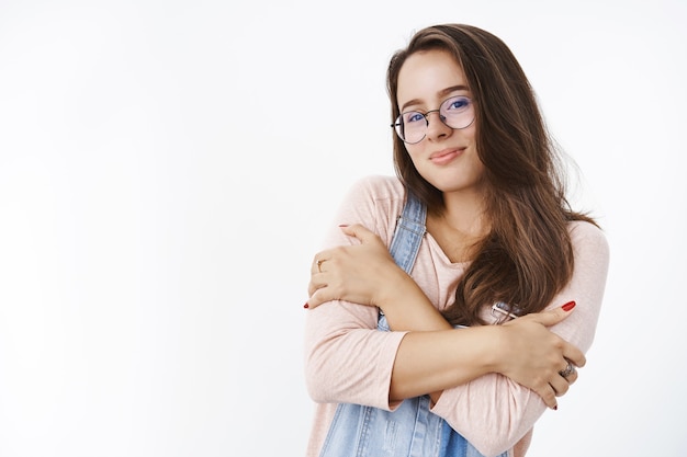 Studio shot of tender silly young brunette in glasses and overalls hugging herself with dreamy pretty smile as if feeling chilly, wanting cuddles in warm hands standing against gray wall.