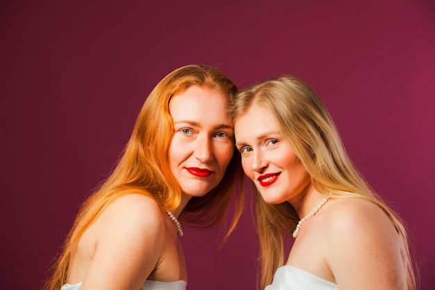 Studio shot of two beautiful sisters blonde and redhead posing in white shirts Two young women with natural beauty on purple background