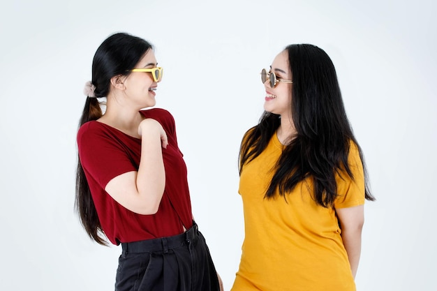 Studio shot of two young Asian cool fashionable stylish female friends in casual tshirt outfit wearing fashion vintage sunglasses standing smiling looking at each other eyes on white background.