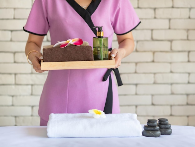 Studio shot unrecognizable Thai professional massaging masseuse in uniform holding body spa treatment stuff herbal compress ball, plumeria flowers, coconut oil and cosmetic on light flare background.