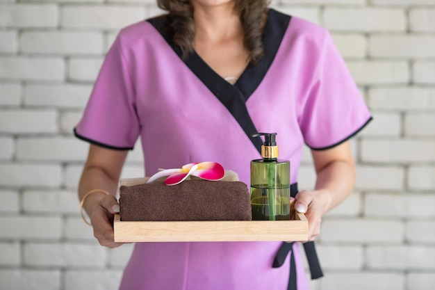 Studio shot unrecognizable Thai professional massaging masseuse in uniform holding body spa treatment stuff herbal compress ball, plumeria flowers, coconut oil and cosmetic on light flare background.