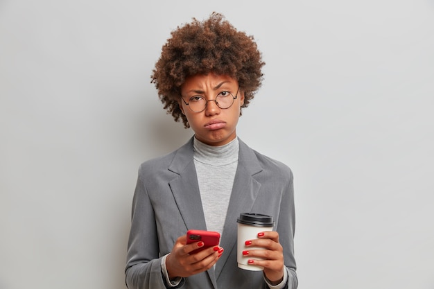 Studio shot of upset fatigue young woman looks with tired expression at front drinks refreshing coffee dressed in formal grey costume uses modern cellular for checking news online stands indoor