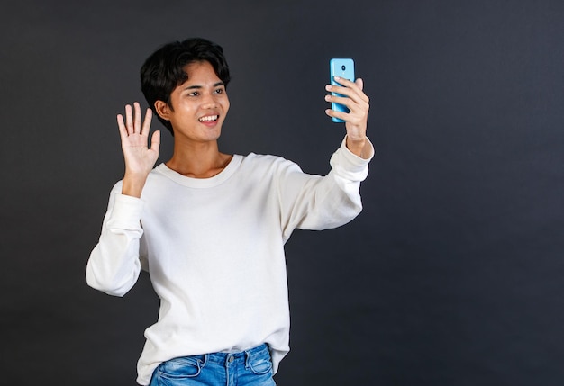 Studio shot van Aziatische jonge Lgbtq homo glamour knappe biseksuele homoseksuele mannelijk model in casual outfit staande glimlachend bedrijf smartphone video-oproep groet zeg hallo hallo vaarwel op zwarte achtergrond