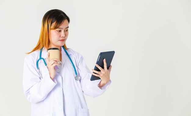 Studio shot van Aziatische succesvolle professionele zelfverzekerde vrouwelijke arts in laboratoriumjas uniforme hangende stethoscoop om nek koffiepauze nemen staande lachende tabletcomputer te houden op witte achtergrond.