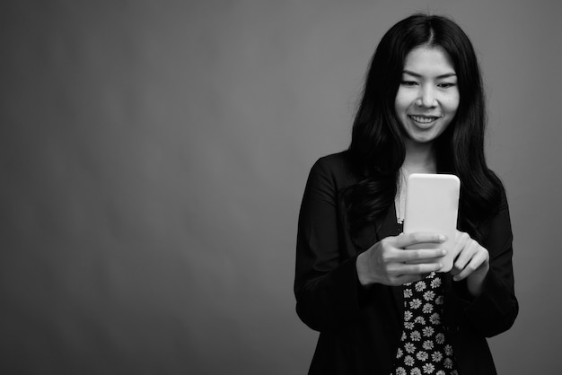 Studio shot van Aziatische vrouw die mobiele telefoon gebruikt tegen een grijze achtergrond in zwart-wit