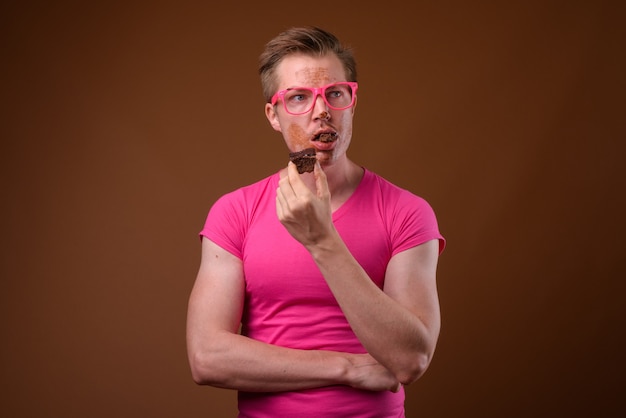 Studio shot van de jonge knappe man met roze shirt met bijpassende roze bril terwijl het hebben van chocoladetaart tegen bruine achtergrond