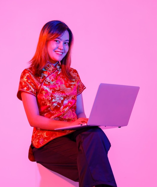 Studio shot van duizendjarige Aziatische jonge tiener vrouwelijke model in mooie rode Chinese traditionele cheongsam qipao shirt zitten met laptopcomputer surfen online winkelen op roze lichte achtergrond.